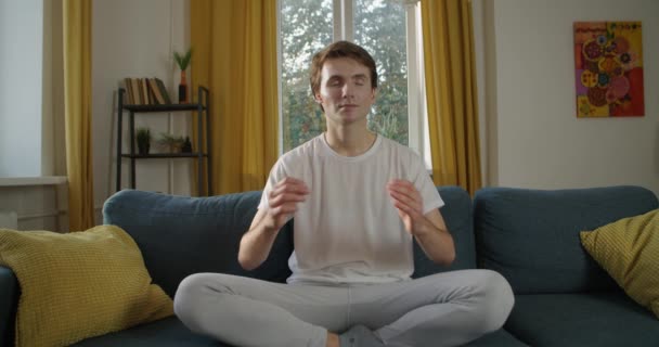 Молодий чоловік медитує вдома з закритими очима в приміщенні, сповільнюючи рух. Передній вид на Кавказького хлопця, який тримає долоні на дошці йоги, розслабляючись на природному світлі дивана. Мирний спокій напружує полегшення — стокове відео