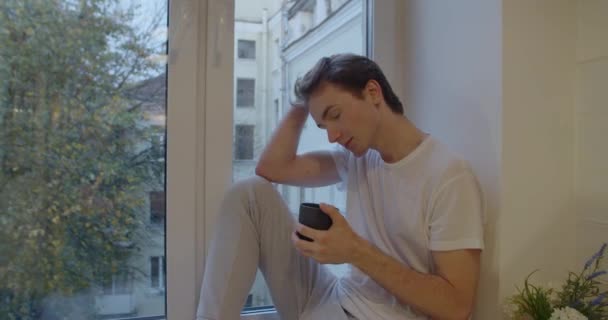 Retrato de mano del hombre pensativo sentado en el alféizar de la ventana con una taza de té en cámara lenta. Vista lateral del tipo bebiendo café por ventana en interiores pensando en problemas de vida. Relaciones conflicto soledad soledad — Vídeos de Stock