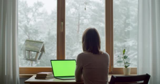 Achteraanzicht van jonge vrouw zittend aan tafel door raam dromen denken aan het leven binnen. Gezichtloos meisje dat thuis aan het typen is op laptop notebook slow motion natuurlijk licht. Thuis blijven concept — Stockvideo