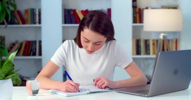Улыбающаяся красивая целеустремленная молодая креативная студентка смотрит на экран ноутбука, смотрит образовательный вебинар на семинаре, пишет заметки на рабочем месте — стоковое видео