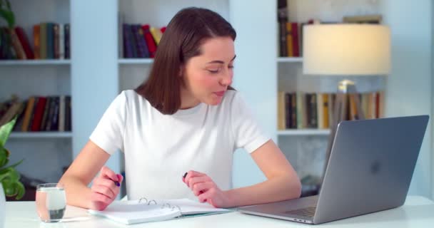 Молодая женщина делает заметки во время работы на ноутбуке в помещении на фоне квартиры. Студентка записывает информацию с компьютера дома в замедленной съемке. Концепция профессии — стоковое видео