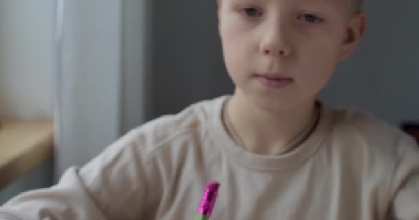 Cerrar retrato frontal de niño mirando lápiz listo para dibujar en el interior luz natural. Niño concentrado haciendo la tarea sentado en la mesa por la ventana en cámara lenta. Concentración educación estancia en casa — Vídeos de Stock