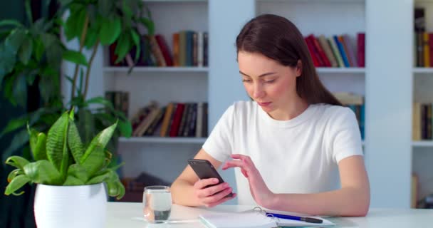 Skoncentrowana kobieta surfująca po Internecie na smartfonie siedzącym przy stole w pomieszczeniach. Młoda studentka pisząca SMS-y na komórce, ucząca się sama w domu. Koncepcja blokady edukacji internetowej — Wideo stockowe