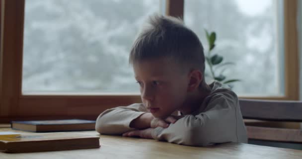 Kimerült iskolás fiú ül a fa asztalnál, és valami lassított felvételen gondolkodik. Közelkép portré szomorú töprengő fiú gyerek nem hajlandó olvasni papírkönyvet a Coronavirus otthoni iskolai oktatás. — Stock videók