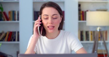 Gülümseyen genç iş kadını akıllı telefonuyla konuşuyor ofis masasında ağır çekimde oturuyor. Serbest çalışan mutlu bir bayan müşteri danışmanı olarak cep telefonu kullanıyor. Web sitesi alışveriş iletişimi