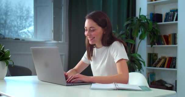 Leende ung kvinna chatta på internet på bärbar dator med glada ansikte inomhus bakgrund. Glada flicka sitter vid bordet skrivbord med hjälp av bärbara dator kommunicera med vänner slow motion. Internetanslutning — Stockvideo