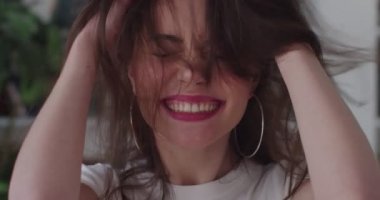 Uzun saçlarını okşayan mutlu stil sahibi kadın içeride ağır çekimde gülüyor. Makyajlı, gülümseyen, titreyen, evde eğlenen bir kadın yüzü. Taze şampuan ürünü. Saç bakım güzellik konsepti