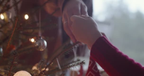 Уютный теплый снимок семейных рук, украшающих елку домашними украшениями ручной работы. Безликая женщина и ребенок висят держа красный орнамент близко к замедленной съемке. праздничные традиции — стоковое видео