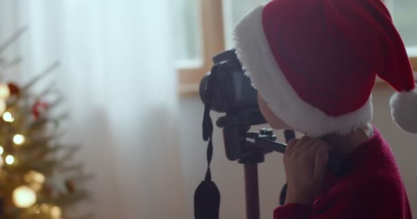 Fiatal gyerek télapó claus kalap lövés karácsonyfa szakmai kamera otthon háttér. Visszapillantás az arctalan fiúra, aki bent fotózott lassított felvételeket készít. Ünnepi memória berendezés technológia — Stock videók