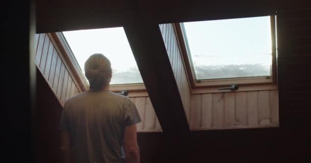 Back view of man standing indoors in wooden interior looking out of window. Faceless person dreaming enjoying sky view planning travel trip for summer. Loneliness covid19 isolation quarantine concept — Stock Video