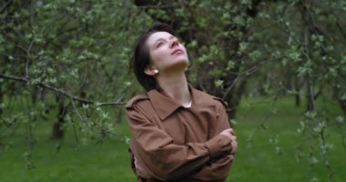 Genç ve şık bir kadın bahar bahçesinde temiz havanın tadını çıkarıyor. Mutluluktan nefes alan yeşil ağaçların arasında tek başına duran hayalperest genç kızın portresi. Yeni yaşam keyfi konsepti