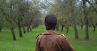 Yeşil ağaçların arasında, yağmurlu havalarda, bahar bahçesinde yürüyen bir kadının arka görüntüsü. Yüzsüz kız dışarıda boş vakit geçirip temiz havanın tadını çıkarıyor. Moda, yalnızlık, yalnızlık, yeni hayat.