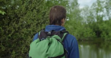 Faceless traveler with backpack standing at lake shore in forest alone sliding shot. Young man tourist has rest while trekking outdoors on nature green background. Travel tourism equipment 