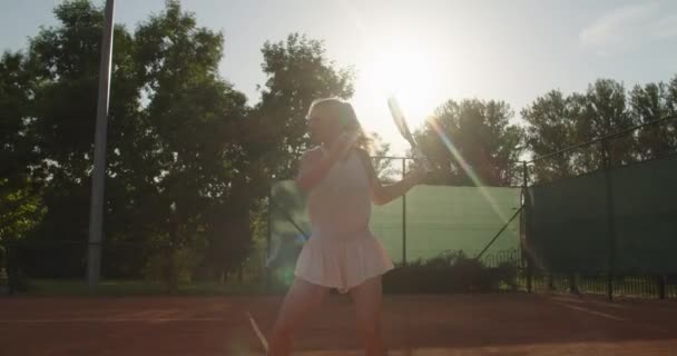Sidovy över ung sexig kvinna slå tennisboll med styrka bakgrundsbelyst med sommarsol. Stark flicka spelare tränar på lera domstol i professionell kostym kläder klänning. Hälsosam livsstil kroppsvård — Stockvideo