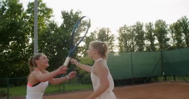 Дві щасливі жінки обіймаються після тенісного матчу, стоячи на глиняному дворі. Обрізати крупним планом вид друзів дівчаток, які займаються спортом разом в літній день. Емоції хобі дозвілля спосіб життя — стокове відео