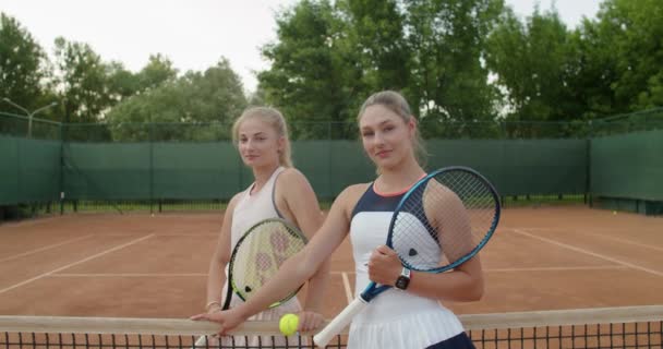 Zoomendes Porträt zweier befreundeter Sportlerinnen, die mit Tennisschlägern vor Sandplatzhintergrund posieren. Ein paar Spielerfrauen, die selbstbewusst und professionell ausgestattet wirken. Aktiver gesunder Lebensstil — Stockvideo