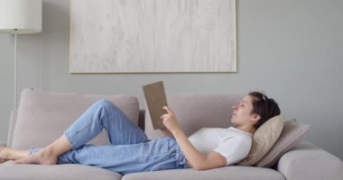 Uykulu kadın sıkı çalışmadan sonra evde rahatlamak için kağıt okuyor. Kanepede uzanmış, uyumaya çalışan kotlu bir kız. Yavaş çekim uzay metni kopyala. Çalışma koşulları kendi kendine izolasyon zamanı