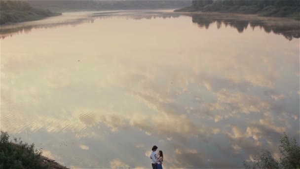 Pareja joven enamorada abrazándose junto al lago al amanecer. Cita de ensueño temprano en la naturaleza. Hermosas nubes se reflejan en las aguas de un lago. Magnífico paisaje acuático con dos personas de pie en la orilla del río. Movimiento lento — Vídeos de Stock