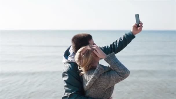 Крупный план молодой счастливой влюбленной пары у моря, позирующей улыбающейся и фотографирующей селфи с помощью камеры смартфона — стоковое видео