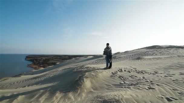 Красивая флайкам-камера с молодой обнимающей парой, стоящей на вершине очаровательного природного песчаного дюнного холма и наслаждающейся видом на море — стоковое видео