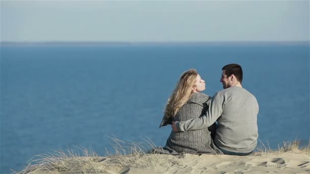 Close up de jovem casal sentado abraçando no topo de uma colina de dunas falando brincando e beijando no fundo do mar azul calmo — Vídeo de Stock