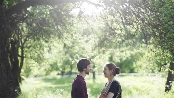 Feliz joven pareja de pie bajo un gran árbol en la naturaleza de verano, hablando y sonriendo. Tiro medio — Vídeo de stock