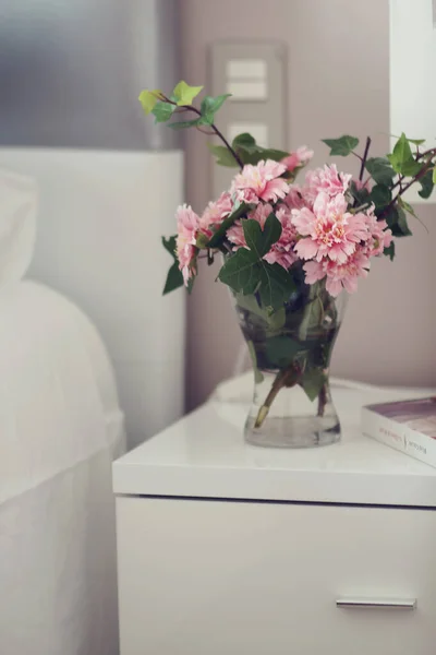 Komodinin yanında pembe çiçekli vazo ve kitapla süslenmiş..