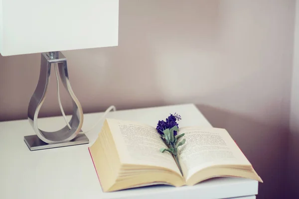 Komodinin yanında pembe çiçekli vazo ve kitapla süslenmiş..