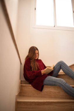 Güzel genç bir bayan kitap okuyor ve mutlu bir şekilde kahve içiyor. Uzun kahverengi saçlı bir kadın kitap okuyor. Merdivenlerde oturan kadın..