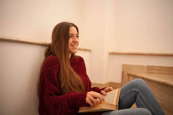 pretty young woman reading a book and drinking coffee happily. Woman with long brown hair reading a book. Woman sitting on some stairs.