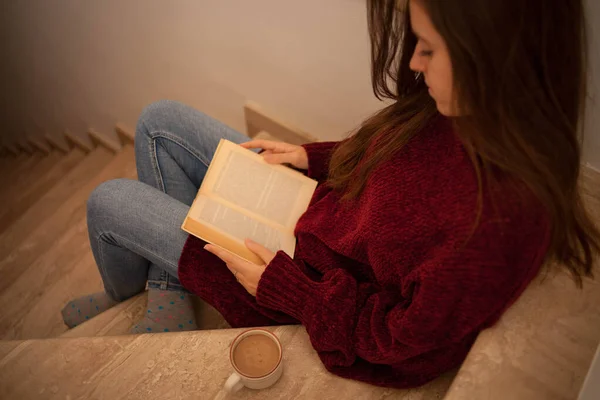 Güzel genç bir bayan kitap okuyor ve mutlu bir şekilde kahve içiyor. Uzun kahverengi saçlı bir kadın kitap okuyor. Merdivenlerde oturan kadın..