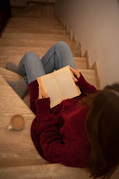 Güzel genç bir bayan kitap okuyor ve mutlu bir şekilde kahve içiyor. Uzun kahverengi saçlı bir kadın kitap okuyor. Merdivenlerde oturan kadın..