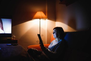 Oturma odasında dokunmatik tablet kullanan yetişkin bir kadın. Yetişkin bir kadın dokunmatik bir cihaz kullanıyor. Kitap okuyan bir kadın. Ailesiyle video görüşmesi yapan bir kadın.