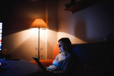 Oturma odasında dokunmatik tablet kullanan yetişkin bir kadın. Yetişkin bir kadın dokunmatik bir cihaz kullanıyor. Kitap okuyan bir kadın. Ailesiyle video görüşmesi yapan bir kadın.