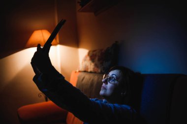 Oturma odasında dokunmatik tablet kullanan yetişkin bir kadın. Yetişkin bir kadın dokunmatik bir cihaz kullanıyor. Kitap okuyan bir kadın. Ailesiyle video görüşmesi yapan bir kadın.