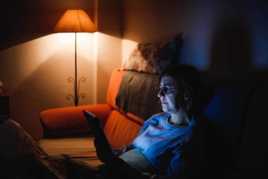 Oturma odasında dokunmatik tablet kullanan yetişkin bir kadın. Yetişkin bir kadın dokunmatik bir cihaz kullanıyor. Kitap okuyan bir kadın. Ailesiyle video görüşmesi yapan bir kadın.