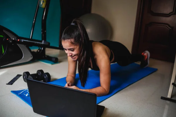 Woman exercising at home with laptop. Fit woman doing yoga plank and watching online tutorials on laptop