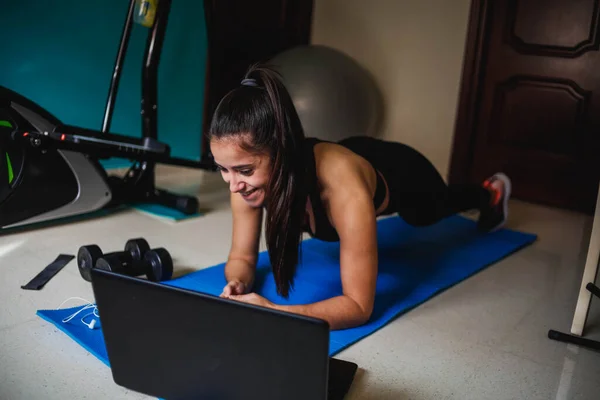 Evde dizüstü bilgisayarla egzersiz yapan bir kadın. Sağlıklı kadın yoga tahtası yapıyor ve dizüstü bilgisayardan online dersler izliyor.
