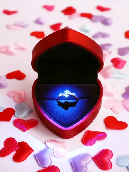 Порпозальная коробка с бриллиантовым ярким кольцом в форме сердца на светлом фоне, украшенном пастельными сердцами. Плакат Дня Святого Валентина. Синий свет на кольце. Гламурное предложение руки и сердца — стоковое фото