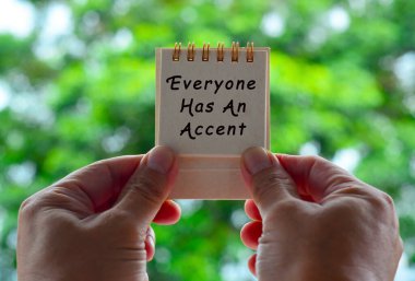 Yazılı ve bulanık yeşil arkaplanı olan beyaz bir not defteri tutan eller. Herkesin bir aksanı vardır.