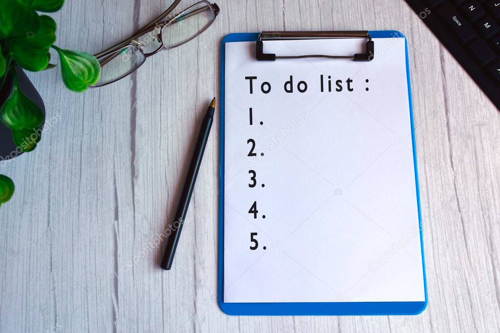 To do list text on blue clip board with glasses, pen, plant and keyboard on wooden desk. 