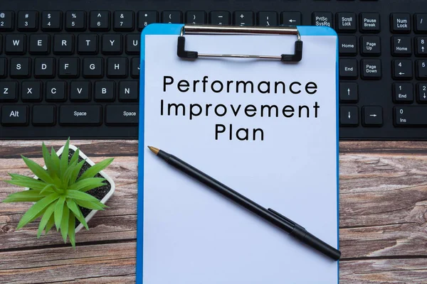 Performance improvement plan text on blue clipboard on top of a keyboard with pen and potted plant on wooden desk