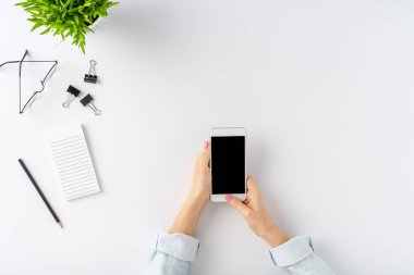 Kadın elleri beyaz masanın üzerinde boş ekranlı cep telefonu kullanıyor. Ofis masaüstü konsepti. Üst görünüm