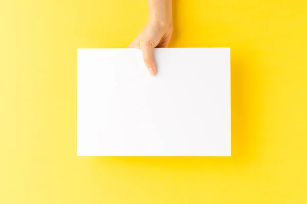 Woman hand showing blank a4 paper sheet on yellow background. Close up