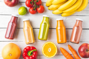 Beyaz ahşap arka planda meyve ve sebzelerle dolu şişelerde çok renkli meyve suları. Üst görünüm