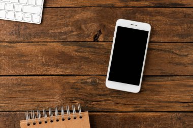 Ahşap arka planda boş ekranlı, bilgisayar klavyesi ve not defteri olan zarif bir akıllı telefon. Ofis masaüstü konsepti