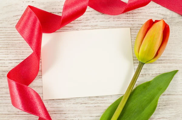 Άδειο καρτ ποστάλ με λουλούδι και κόκκινης κορδέλλας σε ξύλινο υπόβαθρο — Φωτογραφία Αρχείου