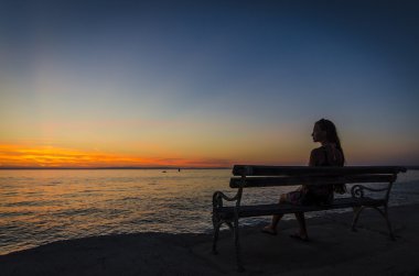Bankta oturan ve gün batımında denizin üzerinde seyir genç kız