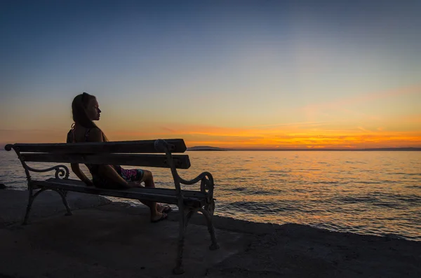 Jovem sentada no banco e olhando para o pôr do sol sobre o mar — Fotografia de Stock