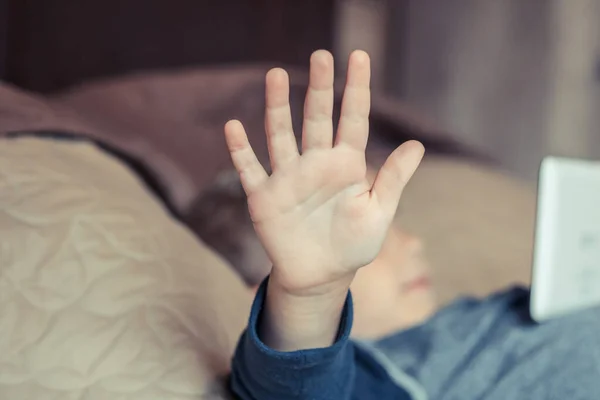 Kid showing stop sign with his hand while lying on the bed and using touchpad.